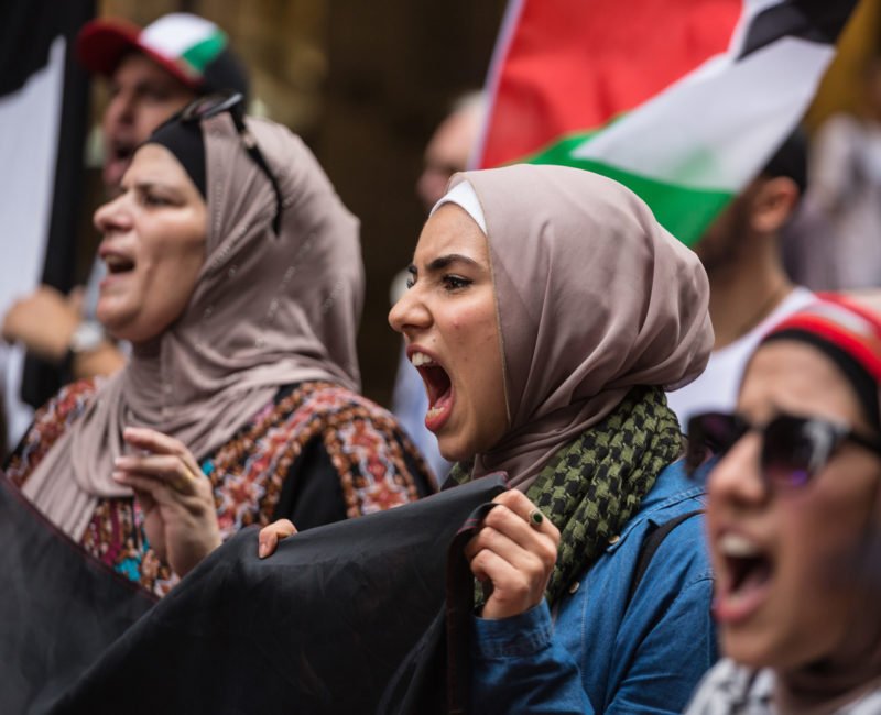 Women in Muslim and traditional Palestinian costume holding a banner and chanting slogans.