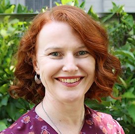A photo of a red-haired woman smiling at the camera, with foliage in the background.