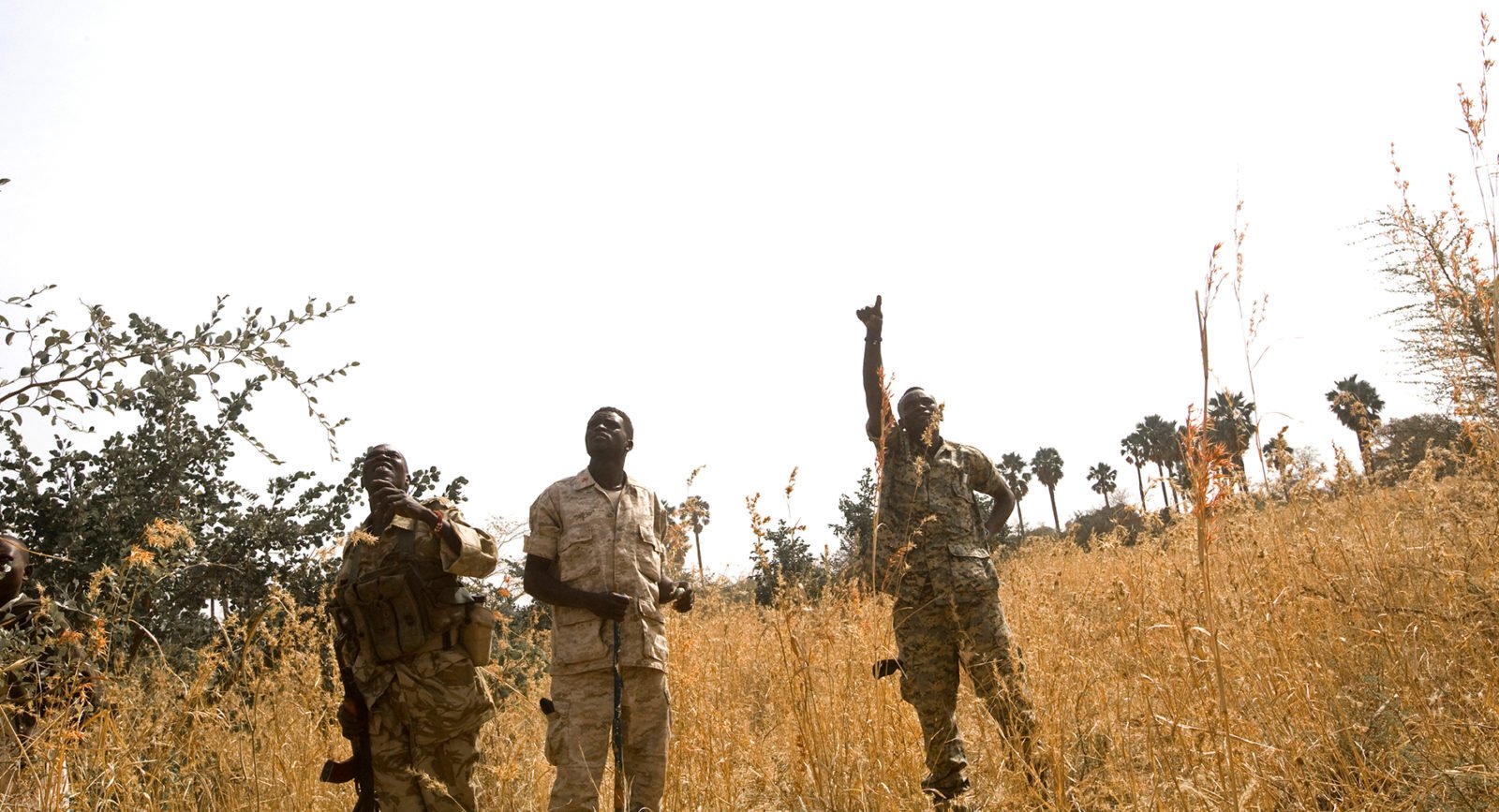 Two military chaplains and a soldier point to the sky as an airplane passes overhead in Sudan