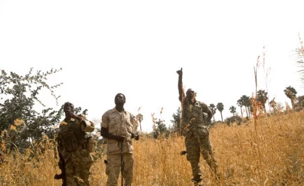 Two military chaplains and a soldier point to the sky as an airplane passes overhead in Sudan