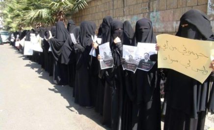 Women wearing black niqab stand in front of a wall holding protest signs in Arabic