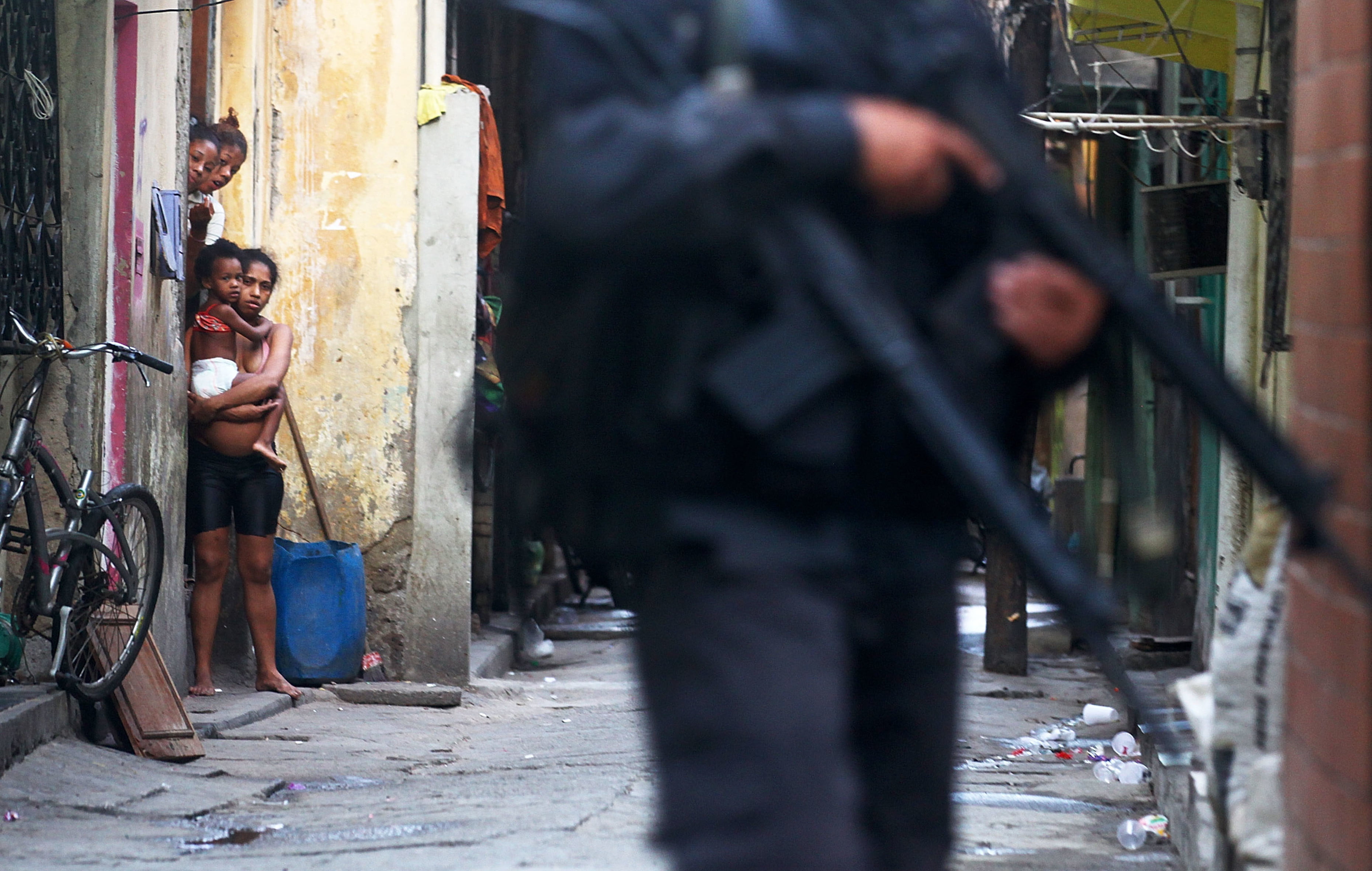 a person in dark police uniform patrols a street with a large gun, onlookers peer from doorway in the background