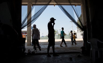 Men stand ouside and under a concrete roof at the Softex refugee camp in Sindos, near Thessaloniki.