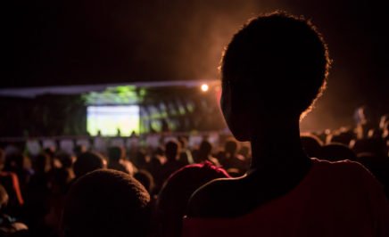 The shadow of a person at the back of a large crows, all facing towards a big screen in the distance