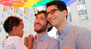 Two men standing together, smiling and holding a young child under a rainbow flag.