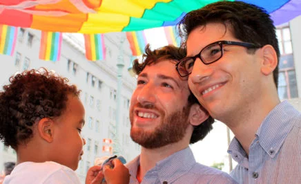 Two men standing together, smiling and holding a young child under a rainbow flag.