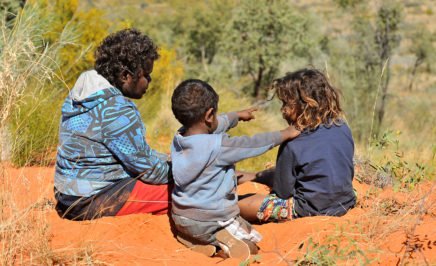 Three young Indigenous people sitting on red outback sand.