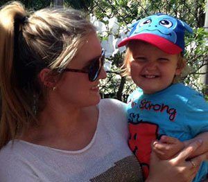 A woman wearing sunglasses looks toward the toddler she is holding, who is smiling and looking at the camera