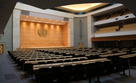 Interior photograph of the large international meeting chamber room at the UN in Geneva, Switzerland. The room is empty.