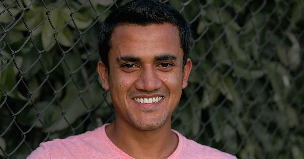 Head and shoulders of Imran, a smiling young man , with a wire fence and foliage behind him