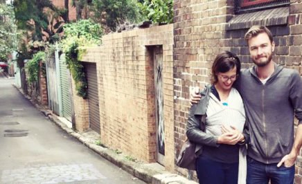 An image of a woman and a man walking down a side street. The man has his arm around the woman and the woman is wearing a newborn baby in a sling.