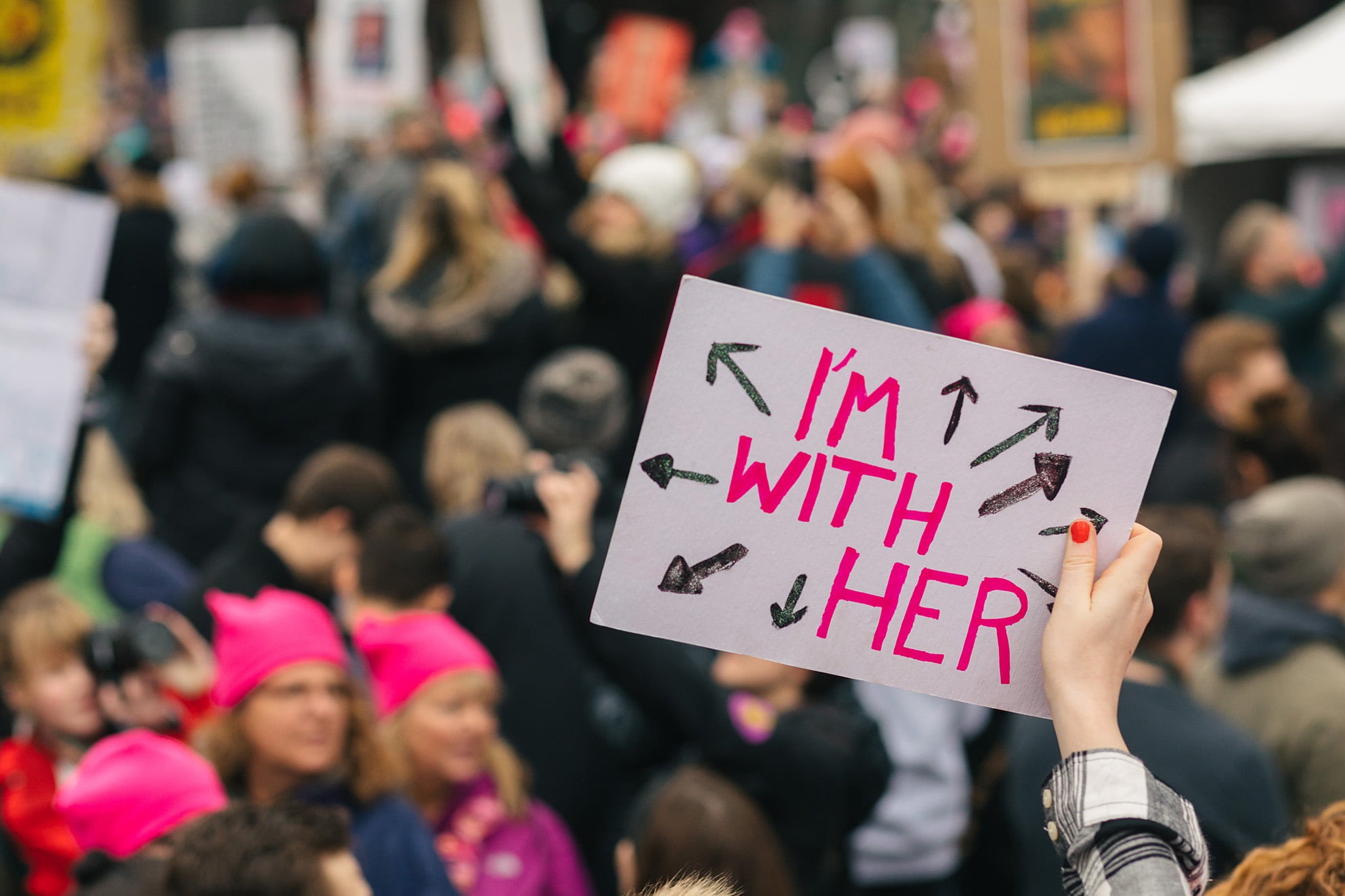 A hand holds up a sign that says "I'm with her", with arrows pointing all around. These arrows point all around at women in the surrounding women's march.