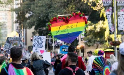 A photo of a crowd of people gather for a rally marriage equality rally in Sydney. There are numerous people holding rainbow flags and carrying signs that say 'Love!' and 'Vote Yes!'