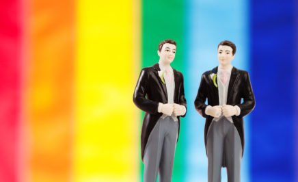 Two groom figures on a wedding cake in front of a pride flag. © iStock/YinYang