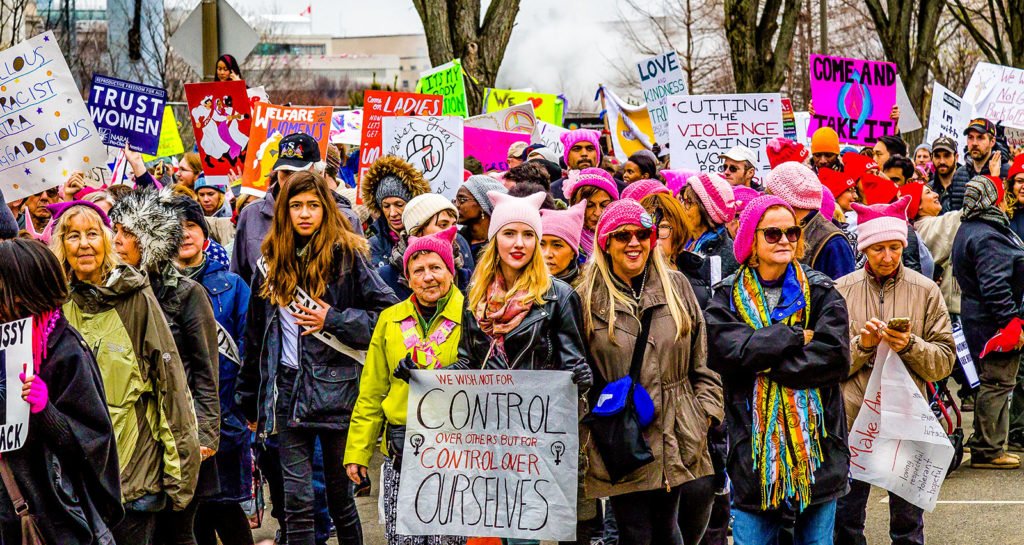 A close up photograph of people taking part in the Women's March on Washington DC on 21 January 2017. The image shows a mixed crowd of men and women holding placards and marching, some of whom are wearing pink hats. A young woman at the front of the shot holds a sign that says 'We wish not for control over others but for control over ourselves.'