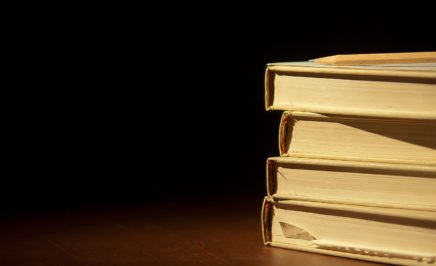 A close up a stack of four books on a wooden table, against a black backdrop.