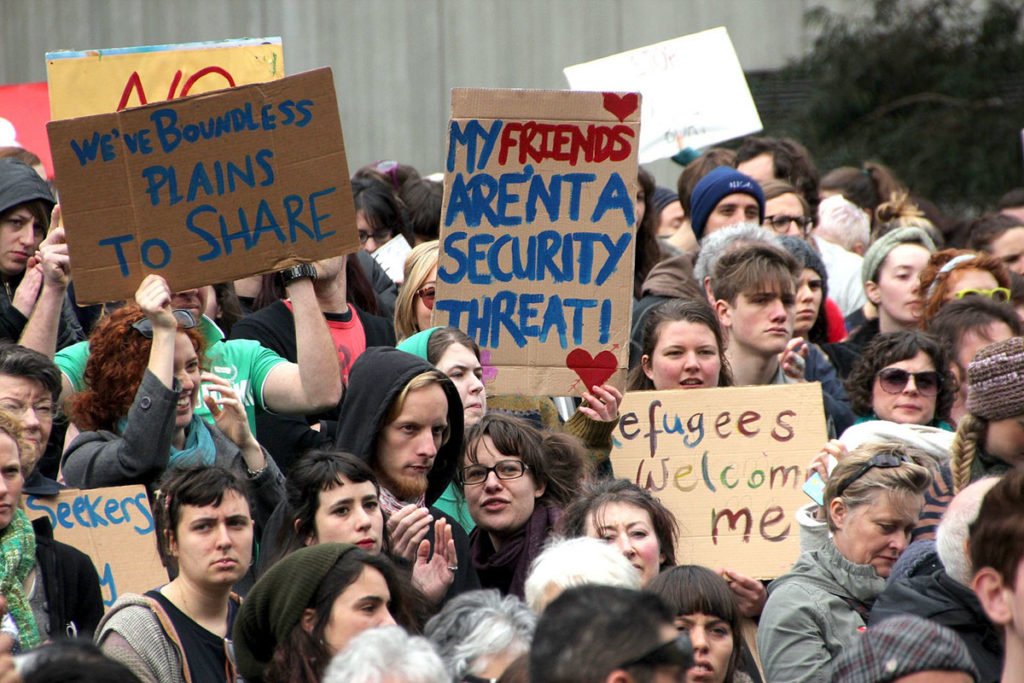 A crowd a rally for refugees. The signs in shot say: 'We've boundless plains to share' and 'My friends aren't a security threat!'