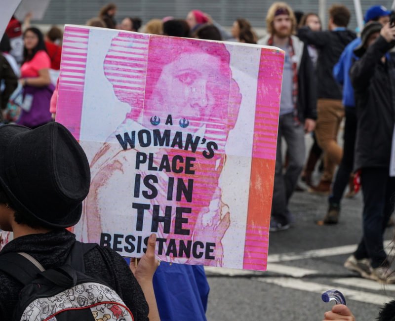 A photograph of a protester in a crowd of people at the women's march on Washington in January 2017. She is holding a sign featuring Carrie Fisher as Princess Leia from Star Wars with the caption 'A women's place is in the resistance'.