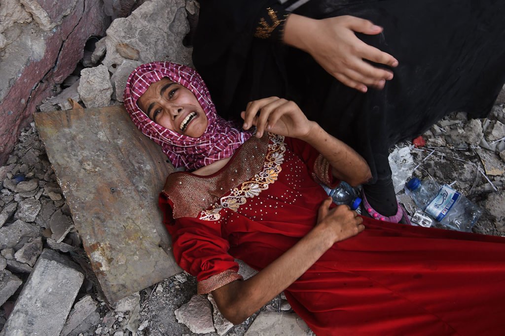 An emaciated teenage girl wearing a red dress and head scarf lies on rubble, an anguished look on her face.