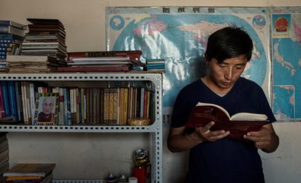 young man reading a book and standing next to a bookcase