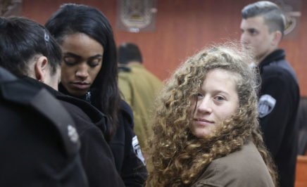 A young woman with curly hair and wearing a brown jacket stands in a court room surrounded by guards. She is looking at the camera.