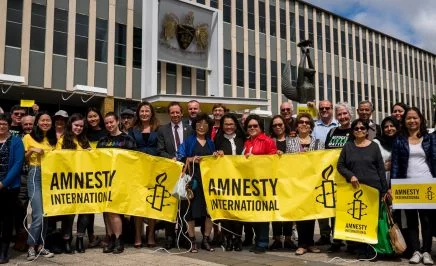 A group of people holding yellow Amnesty banners standing outside Parliament House in Canberra