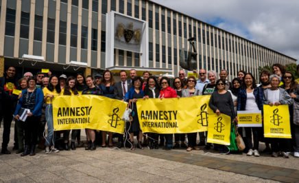 A group of people holding yellow Amnesty banners standing outside Parliament House in Canberra