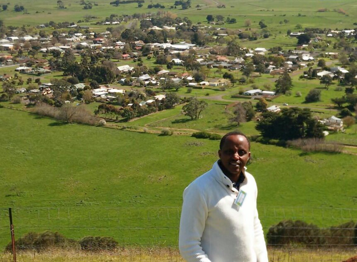 Man with dark skin in business attire with green, rural farmlands behind him.