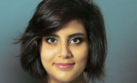 Loujain al-Hathloul smiling into the camera. She is wearing a white top and a necklace and the background behind her is blue