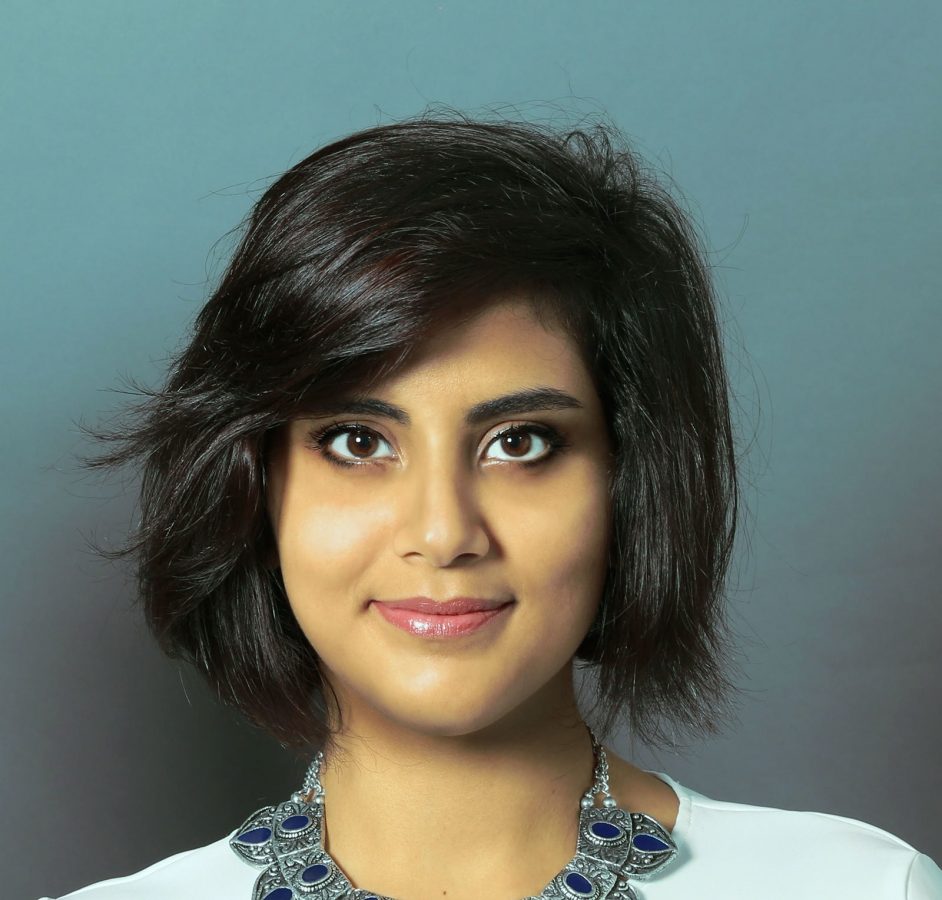 Loujain al-Hathloul smiling into the camera. She is wearing a white top and a necklace and the background behind her is blue