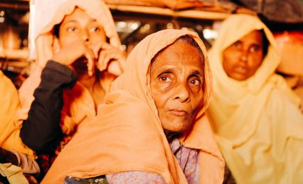 Three Rohingya refugee women staring into the camera. An older woman sits in front of two younger women.