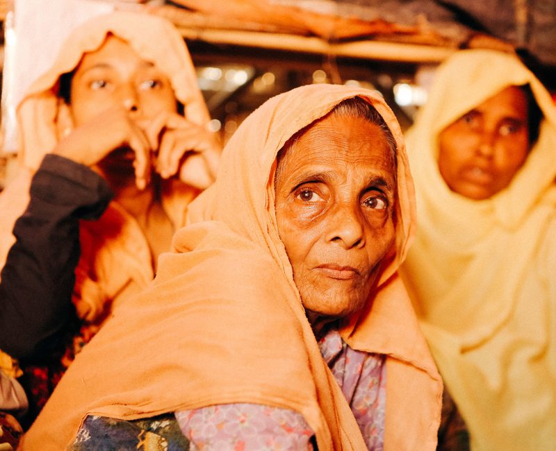 Three Rohingya refugee women staring into the camera. An older woman sits in front of two younger women.