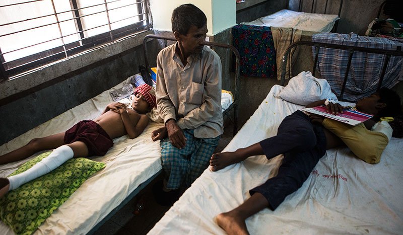 Two Rohingya children lie on hospital beds with bandages on their wounds. A man is watching over the children.