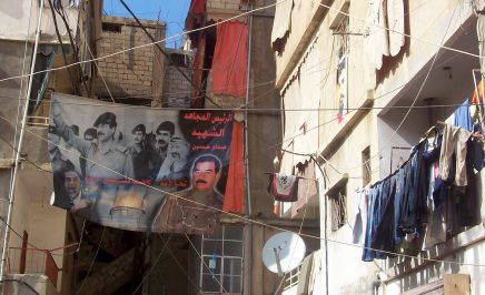 Image of a refugee camp: makeshift buildings and a heavy net of electrical wires crisscrossing between buildings. A sign of with Saddam Hussein's image on it is strung up between buildings. All set against a narrow strip of blue sky.