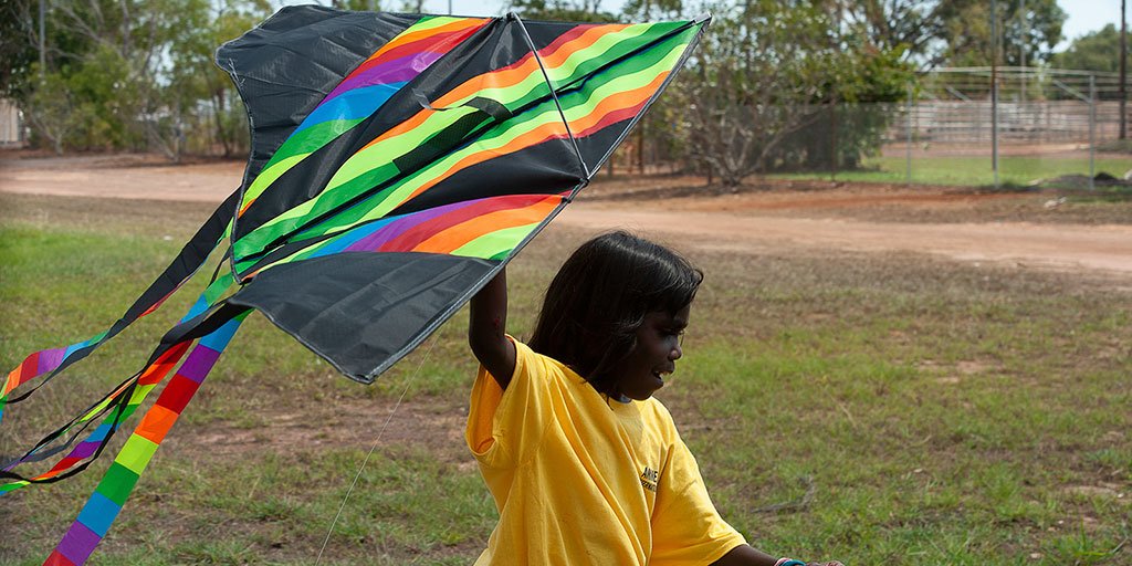 A young girl wearing a yellow t-shirt runs through a field flying a brightly coloured kite above her.