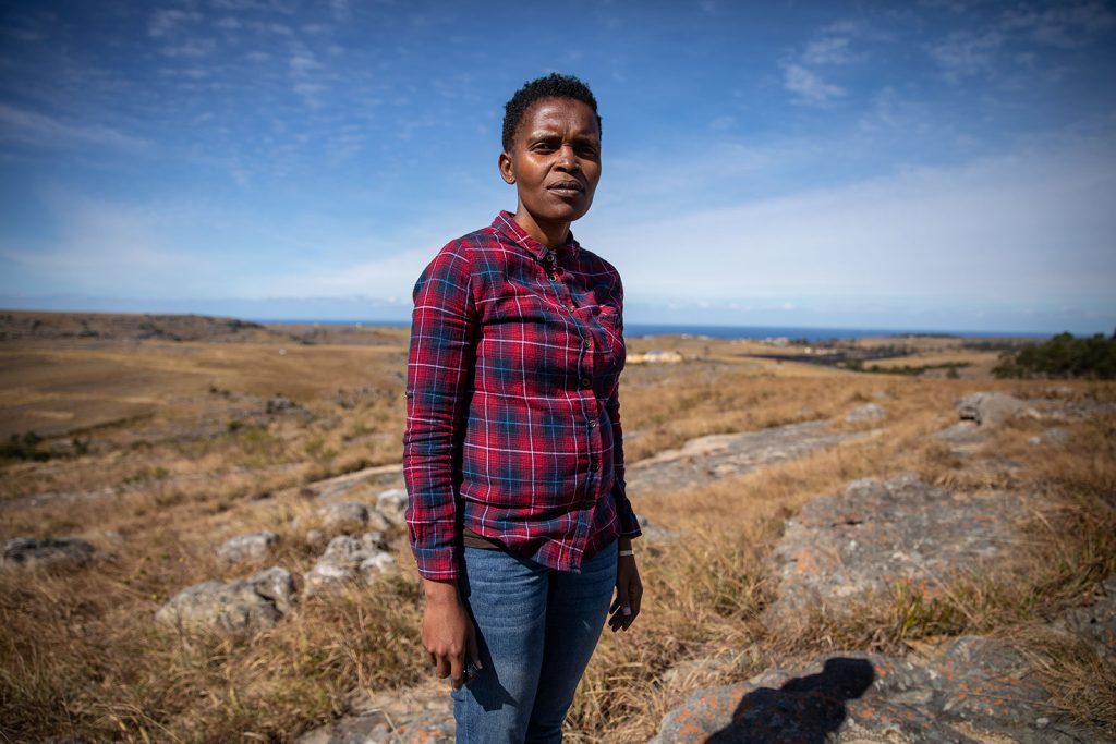 A woman with short hair and wearing jeans and a red and blue checked shirt stands in a field of rocks and grass in the Xolobeni area near Mbizana in Eastern Cape.