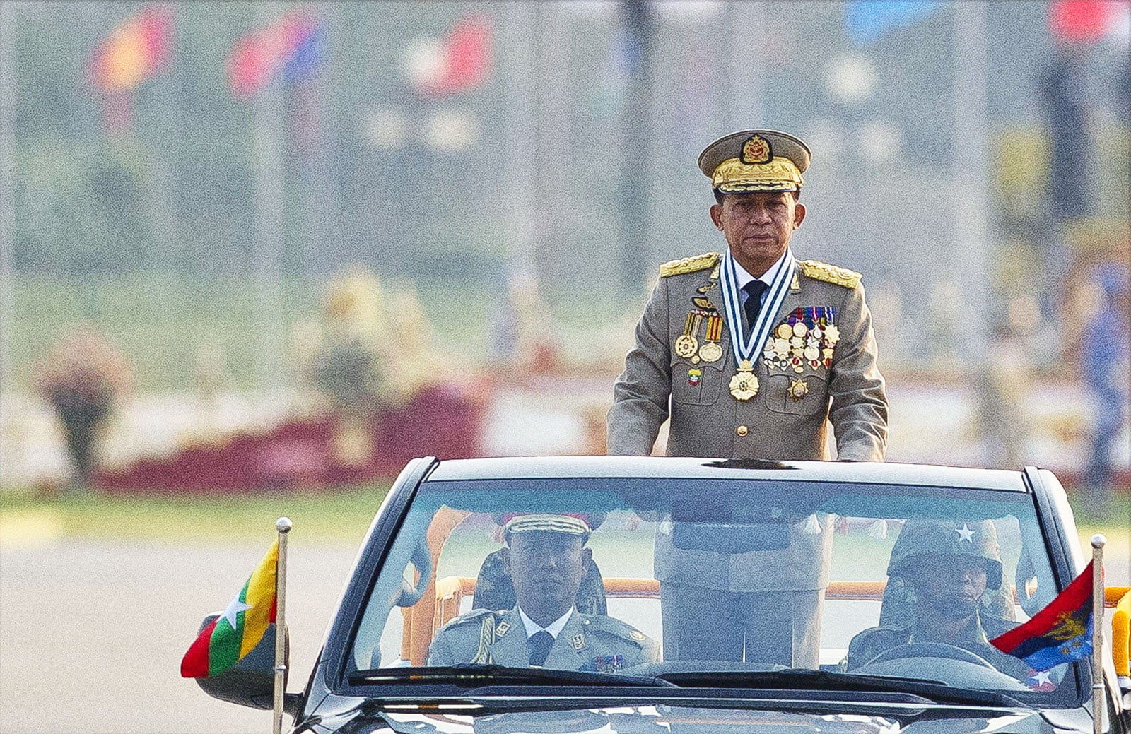 A man in a Myanmar military uniform stands up in an open-top car at a military parade. There are two other people in the car with him, also in military dress. Blurred in the background we can see a line of flags from different countries.