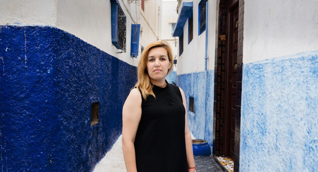 A woman wearing a black top stands in an alleyway painted white and blue and stares towards the camera.
