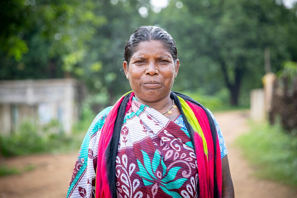 An Indian woman in a brightly coloured sara and scarf smiles at the camera. She is standing close to the camera and behind her we can see the blurred outline of a small building set against a backdrop of leafy green trees.