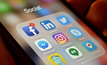 A hand holding a mobile phone displaying the icons of social media apps: Facebook, LinkedIn, Twitter, Messenger, Instagram, Skype, Timehop, Hangouts and Periscope.