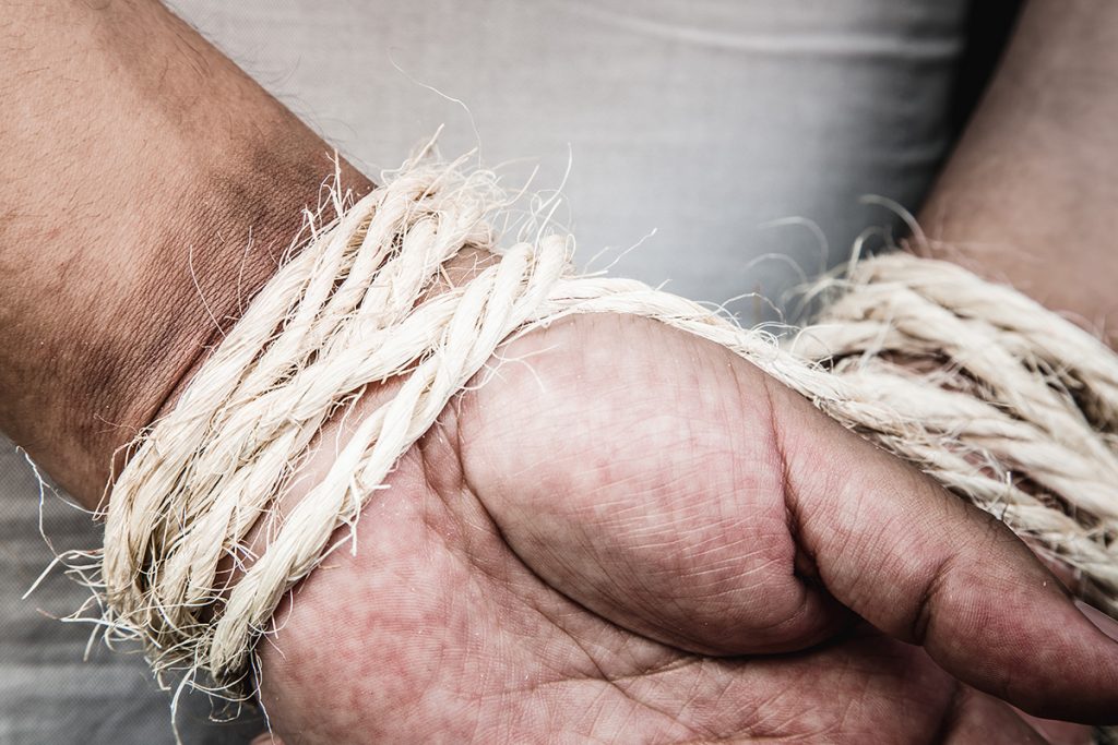 A photo of a man's hands bound behind his back with fraying twine or rope.