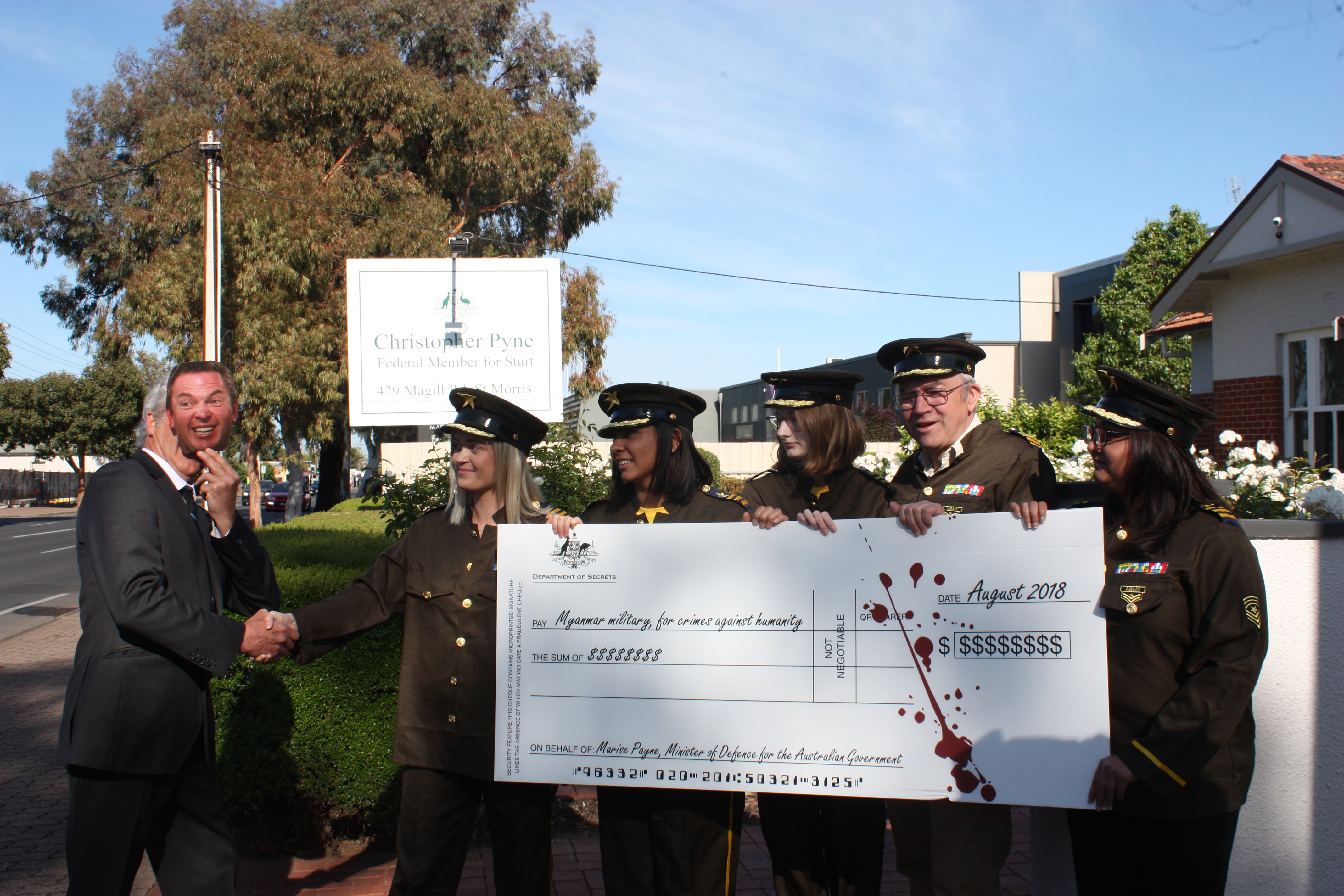 Man with paper mask of Christopher Pyne's face giving large novelty cheque to people dressed in military uniforms