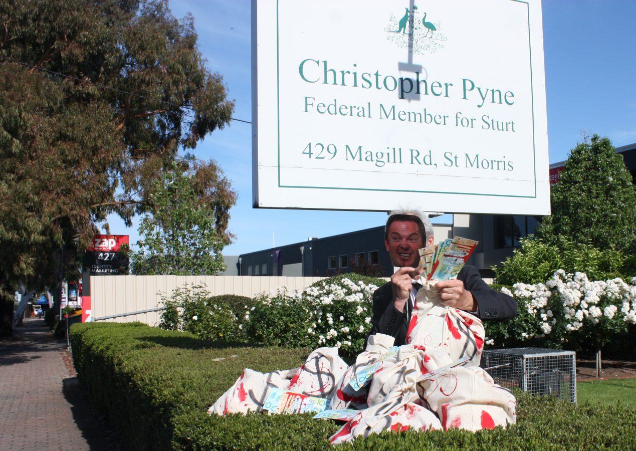 man sits in front of sign saying 'christopher pyne federal member for sturt' with a paper mask, holding bags of money