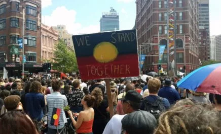 People gather to protest for Indigenous rights. Someone holds a sign that says: 'We stand together'.