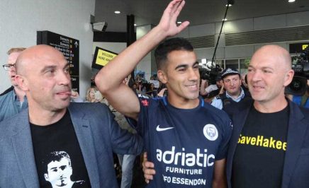 Hakeem in a crowd of media and supporters, smiling and raising his hand in greeting