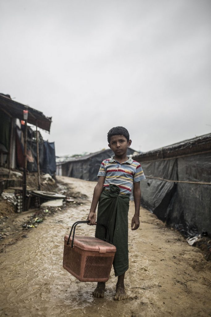 A young boy stands on a muddy path between makeshift refugee tents and looks into the camera. He is carrying a basket.