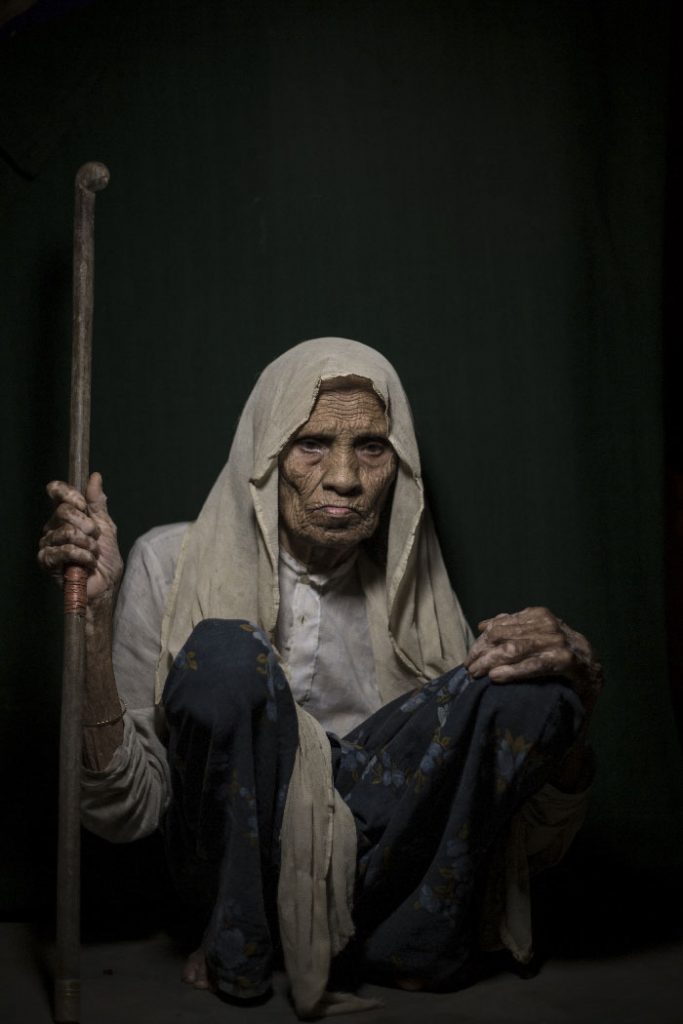 An elderly woman holding a walking stick sits on the floor looking towards the camera.