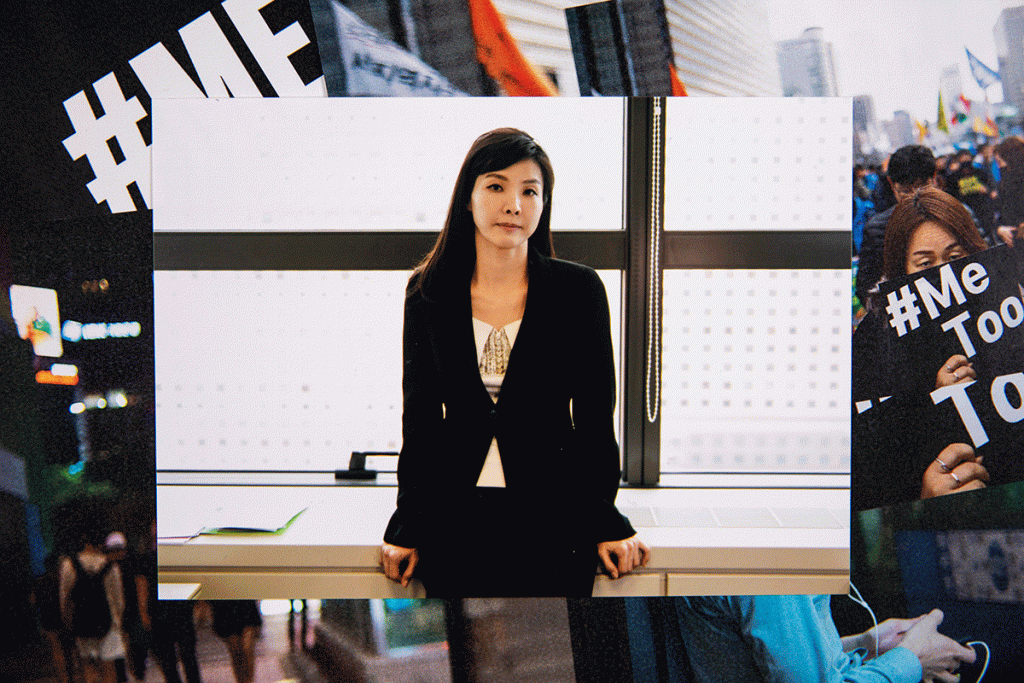 A young South Korean woman leaning against a desk looks towards the camera with a serious expression on her face.