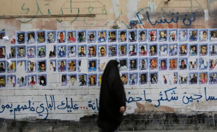 A woman wearing a black headscarf walks past photos of people pasted to an old building's wall, with crumbling plaster. Above and below is spray-painted Arabic words