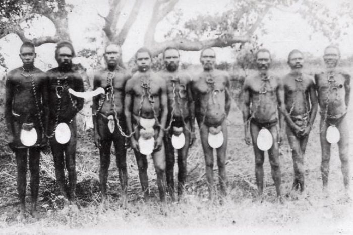 Black and white blurry image of Aboriginal people stand in a row, chained together by their necks, wearing riji (carved pearlshell) as they stand in the mangroves of Broome, c. 1910.